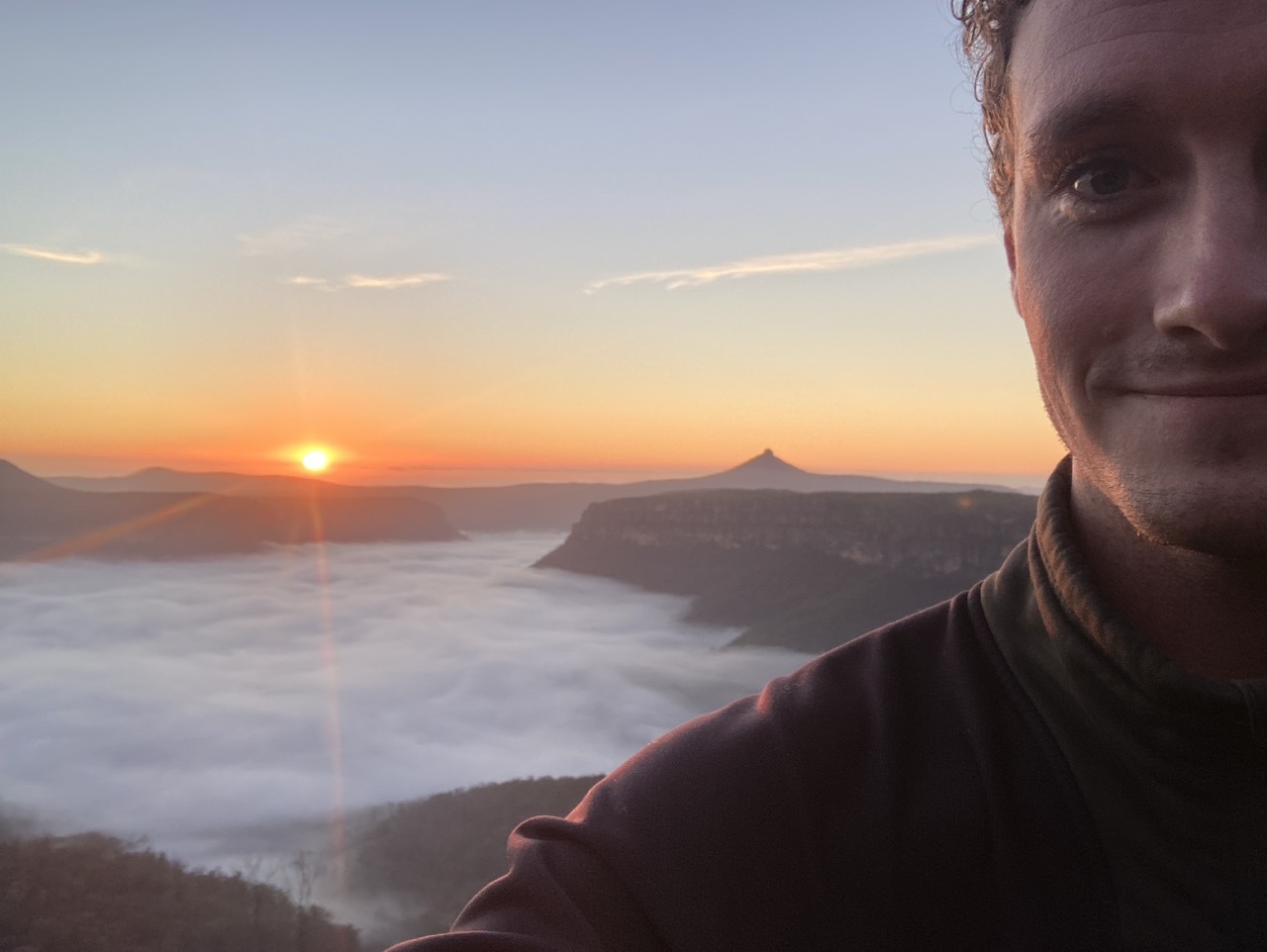 A person takes a selfie at sunrise with a scenic view of mountains and a valley covered in mist or low clouds, with the sun rising on the horizon.