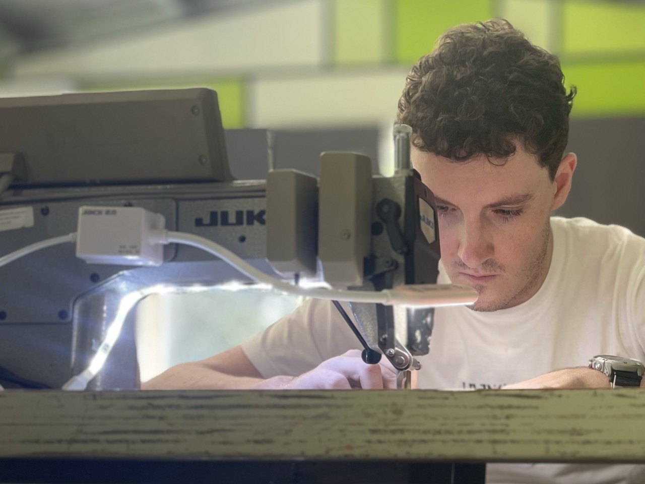 A man concentrates while sewing fabric with an industrial JUKI sewing machine in a well-lit workspace, with green and gray panels in the background.